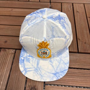 May include: A white baseball cap with a blue marble effect design. The cap has a curved peak and a gold emblem with the text "LONDON POLICE ASSOC". The cap is set against a wooden background.