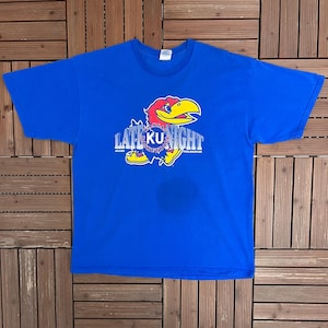 May include: A royal blue t-shirt with a cartoon Jayhawk mascot graphic and the text "LATE KU NIGHT". The Jayhawk is red, yellow, and blue. The text is white and blue. The shirt is displayed on a wooden surface.