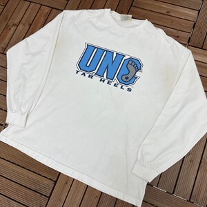 May include: White long-sleeved t-shirt with the text "UNC TAR HEELS" in blue and grey. The shirt has a crew neck and long sleeves. The graphic includes a stylized foot. The shirt is made of a soft material.