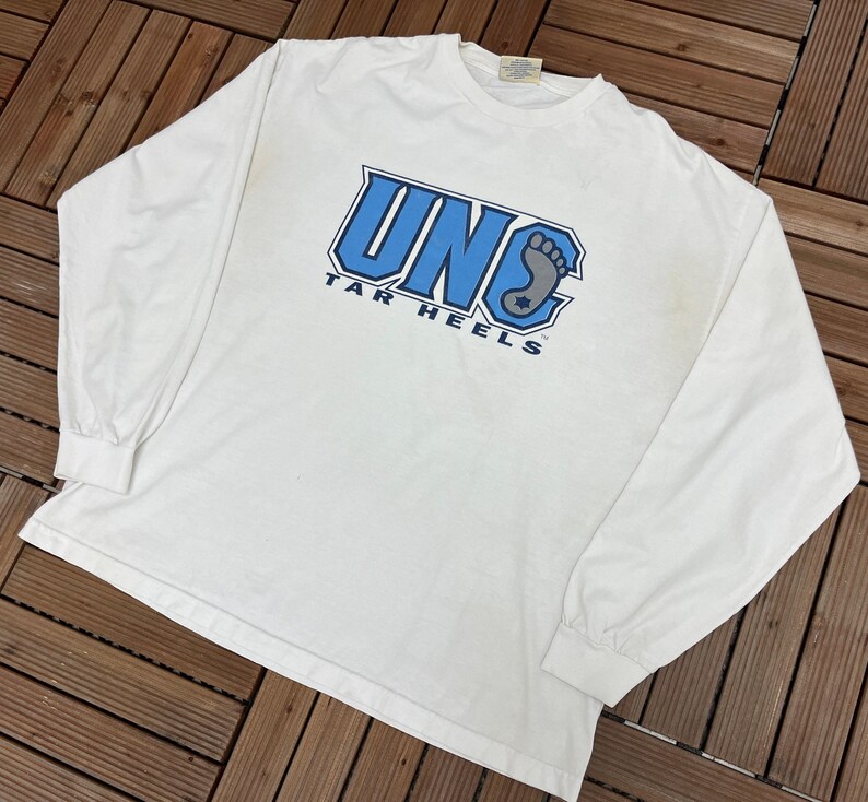 May include: White long-sleeved t-shirt with the text "UNC TAR HEELS" in blue and grey. The shirt has a crew neck and long sleeves. The graphic includes a stylized foot. The shirt is made of a soft material.