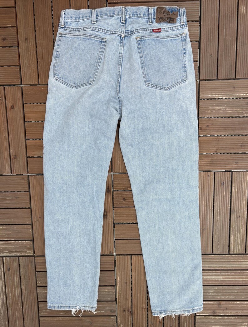 May include: Light blue denim jeans with a classic five-pocket design. The jeans have a faded wash and a straight leg fit. The back pockets are visible, and the Wrangler brand patch is at the waistband.