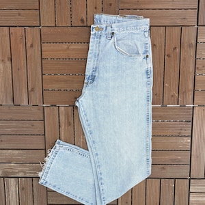 May include: Light blue denim jeans with a classic five-pocket design. The jeans have a button and zipper closure, belt loops, and a straight leg cut. The denim has a faded wash, giving it a worn-in look. The jeans are folded on a wooden surface.