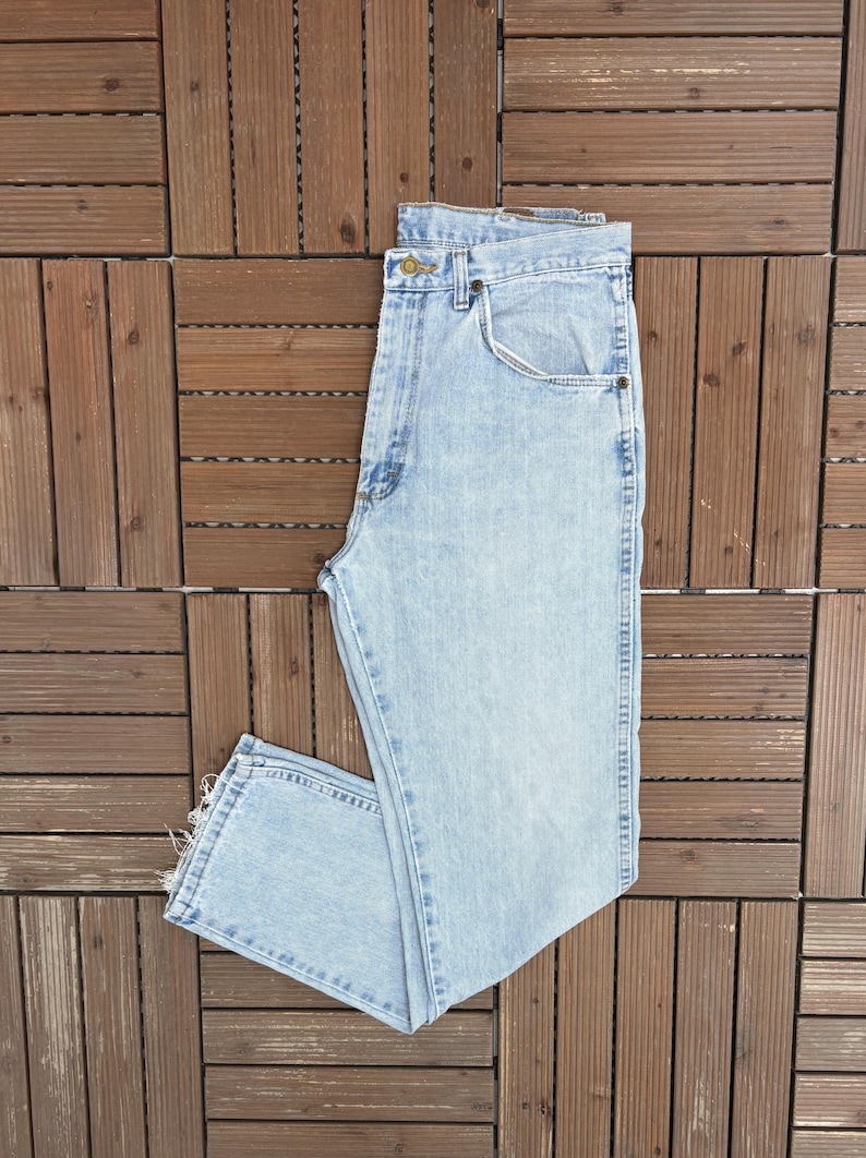 May include: Light blue denim jeans with a classic five-pocket design. The jeans have a button and zipper closure, belt loops, and a straight leg cut. The denim has a faded wash, giving it a worn-in look. The jeans are folded on a wooden surface.