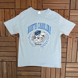 May include: Light blue t-shirt featuring a graphic of a ram with the text "North Carolina Tar Heels" in blue. The design is encircled by a laurel wreath. The shirt has short sleeves and a crew neck.