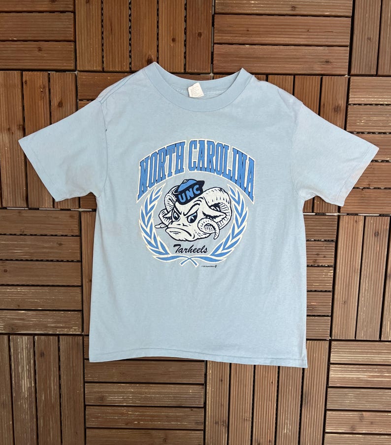 May include: Light blue t-shirt featuring a graphic of a ram with the text "North Carolina Tar Heels" in blue. The design is encircled by a laurel wreath. The shirt has short sleeves and a crew neck.