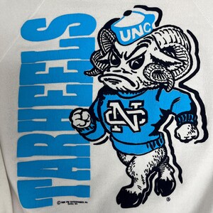 K&ouml;nnte beinhalten: Cremefarbenes Sweatshirt mit einem blauen Cartoon-Widder-Maskottchen, das einen Hut mit "UNC" und einen blauen Pullover tr&auml;gt. Das Wort "TARHEELS" ist vertikal in Blau aufgedruckt. Das Sweatshirt hat einen Vintage-Look.