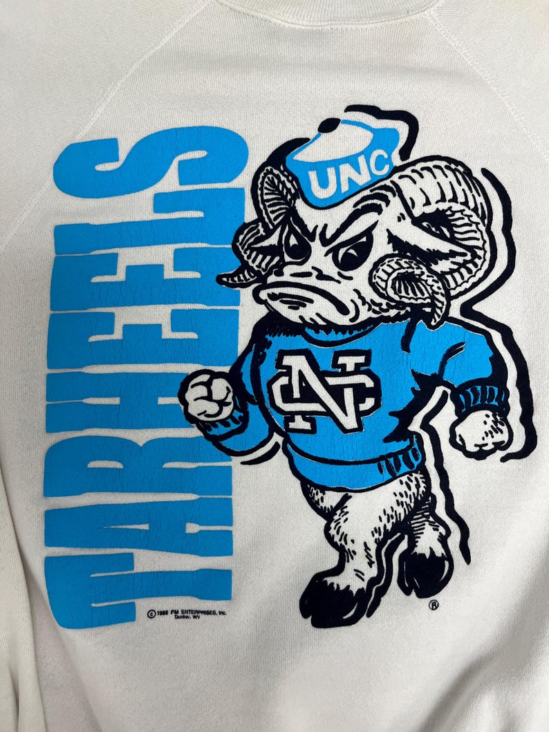 K&ouml;nnte beinhalten: Cremefarbenes Sweatshirt mit einem blauen Cartoon-Widder-Maskottchen, das einen Hut mit "UNC" und einen blauen Pullover tr&auml;gt. Das Wort "TARHEELS" ist vertikal in Blau aufgedruckt. Das Sweatshirt hat einen Vintage-Look.