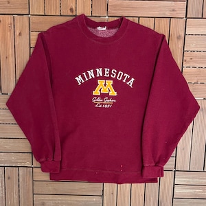 May include: A maroon crewneck sweatshirt featuring the word MINNESOTA in white arched lettering above a yellow M logo. Below the logo, the text reads Golden Gophers and Est. 1851 in white script and print. The garment has long sleeves and ribbed cuffs.