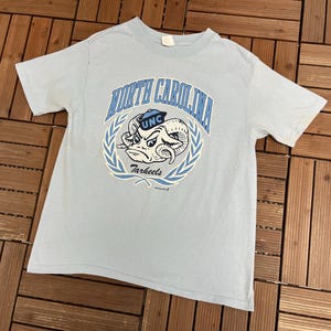 May include: Light blue t-shirt featuring a graphic of a ram with the text "North Carolina" and "Tarheels" in a circular design. The shirt has short sleeves and a crew neck.