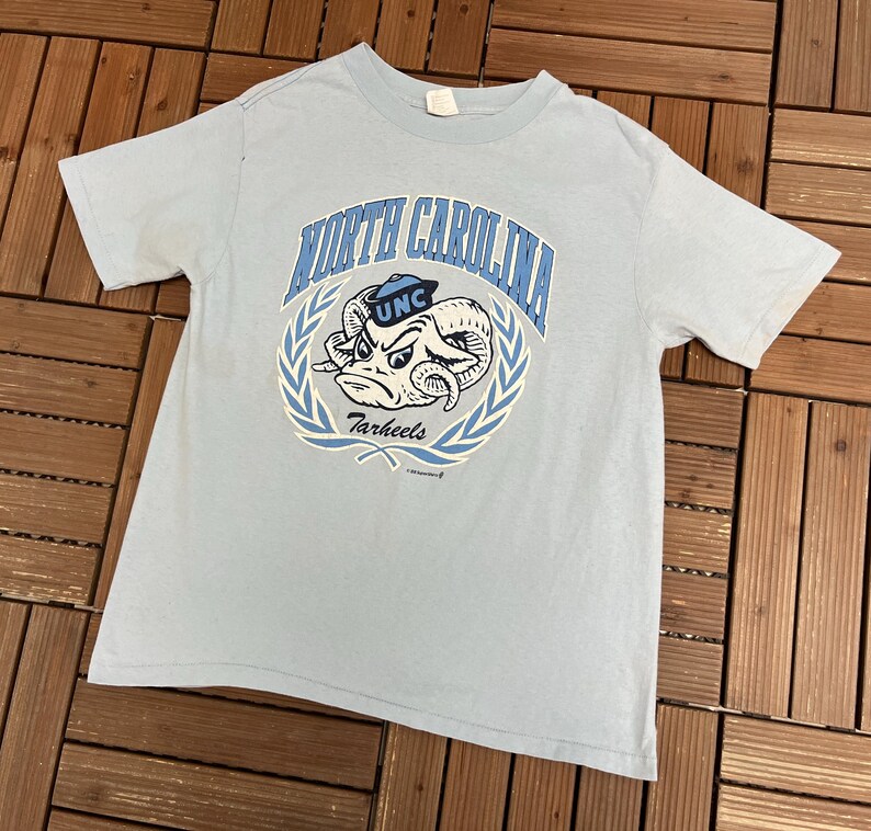 May include: Light blue t-shirt featuring a graphic of a ram with the text "North Carolina" and "Tarheels" in a circular design. The shirt has short sleeves and a crew neck.