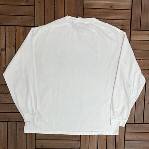 May include: A white long-sleeved top with a crew neck. The shirt features long sleeves with ribbed cuffs. The garment is made of a soft, opaque fabric. The shirt is laid flat on a wooden surface.
