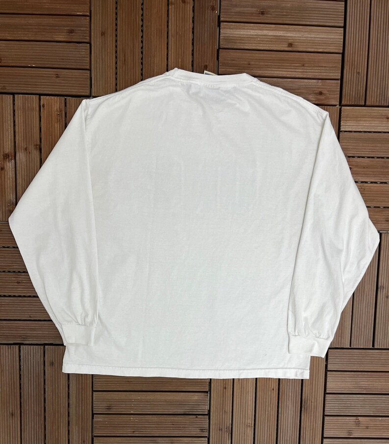 May include: A white long-sleeved top with a crew neck. The shirt features long sleeves with ribbed cuffs. The garment is made of a soft, opaque fabric. The shirt is laid flat on a wooden surface.