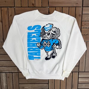 K&ouml;nnte beinhalten: Wei&szlig;es Sweatshirt mit blauem Text "TAR HEELS". Das Sweatshirt zeigt ein Cartoon-Widder-Maskottchen, das ein blaues Trikot mit "UNC" tr&auml;gt. Der Widder befindet sich in Kampfstellung, ein Design f&uuml;r Fans.