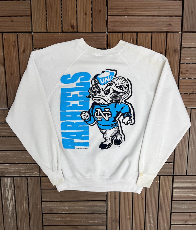 K&ouml;nnte beinhalten: Wei&szlig;es Sweatshirt mit blauem Text "TAR HEELS". Das Sweatshirt zeigt ein Cartoon-Widder-Maskottchen, das ein blaues Trikot mit "UNC" tr&auml;gt. Der Widder befindet sich in Kampfstellung, ein Design f&uuml;r Fans.