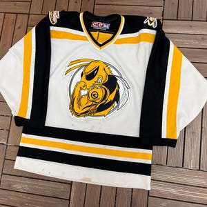 May include: White hockey jersey with black and yellow accents. The jersey features a large graphic of a yellow and black hornet on the front. The sleeves are black with yellow stripes. The jersey has a V-neck collar.