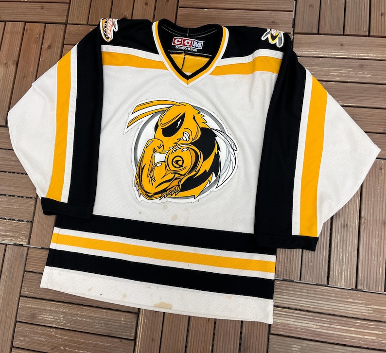May include: White hockey jersey with black and yellow accents. The jersey features a large graphic of a yellow and black hornet on the front. The sleeves are black with yellow stripes. The jersey has a V-neck collar.