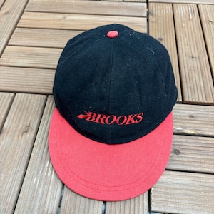 Könnte beinhalten: Eine schwarze Baseballkappe mit rotem Schirm und einem roten Knopf auf der Oberseite. Das Wort BROOKS ist in rotem Garn auf die Vorderseite der Kappe gestickt und enthält ein stilisiertes grafisches Element im ersten Buchstaben des Markennamens.
