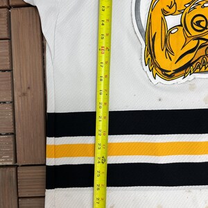 May include: White hockey jersey with black and gold stripes. Features a large yellow and black logo on the upper right. The jersey is approximately 29 inches long, measured with a yellow tape measure.