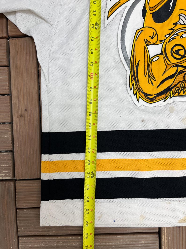 May include: White hockey jersey with black and gold stripes. Features a large yellow and black logo on the upper right. The jersey is approximately 29 inches long, measured with a yellow tape measure.