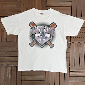 May include: White t-shirt featuring the Arizona Diamondbacks logo. The logo includes the team name, a baseball diamond, and crossed bats in shades of purple, teal, and orange. The shirt is made of a soft material.