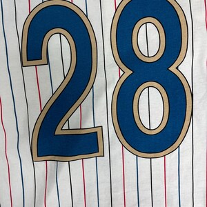 May include: A white fabric garment featuring a repeating pattern of thin vertical pinstripes in alternating red, blue, and black colors. The number 28 is printed in large, bold blue digits with a tan outline centered on the fabric surface.