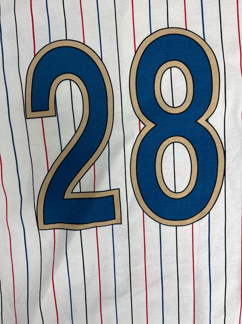 May include: A white fabric garment featuring a repeating pattern of thin vertical pinstripes in alternating red, blue, and black colors. The number 28 is printed in large, bold blue digits with a tan outline centered on the fabric surface.