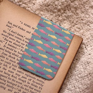 Op de afbeelding: Een rechthoekige boekenlegger met een turquoise achtergrond en een herhalend patroon van kleurrijke vissen. De vissen zijn in tinten geel, roze en paars. De boekenlegger ligt op een open boek.