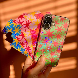 May include: A clear smartphone case featuring a green background with a repeating pattern of pink holographic stars. Behind the phone case, a fan of four additional adhesive stickers displays similar star patterns in blue, pink, orange, and yellow colourways.