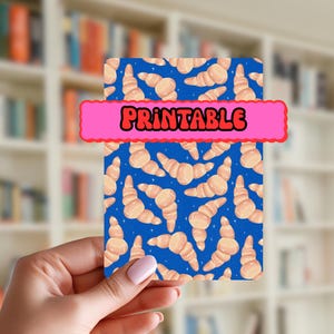 Könnte beinhalten: Eine rechteckige Karte mit blauem Hintergrund und einem Muster aus Croissants. Ein pinkfarbenes Banner mit dem Wort "PRINTABLE" in Weiß und Rot verläuft über die Oberseite der Karte. Die Karte wird in einer Hand gehalten.