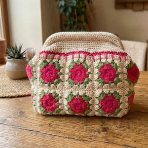 May include: A crocheted handbag with a beige top and a floral pattern in pink and green. The bag is sitting on a wooden table. The bag has a rounded shape and a top closure.
