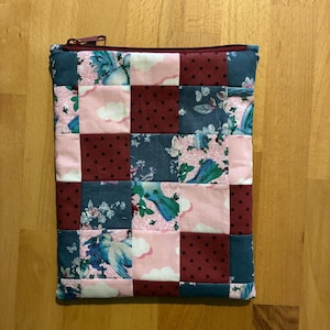 Könnte beinhalten: Eine rechteckige Reißverschlusstasche mit einem Patchwork-Design aus Stoffquadraten. Das Muster besteht aus abwechselnden Blöcken in Dunkelrot mit schwarzen Punkten, einfarbigem Dunkeltürkis und hellrosa Stoff mit weißen Wolken und blauen Vögeln.
