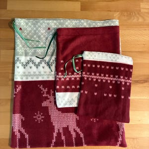 Könnte beinhalten: Drei Stoffbeutel mit Kordelzug in verschiedenen Größen mit einem kastanienbraunen und weißen Muster aus Rentiersilhouetten und geometrischen Formen. Jeder Beutel ist mit einem dünnen grünen Band versehen und hat eine weiche, fleeceartige Textur.