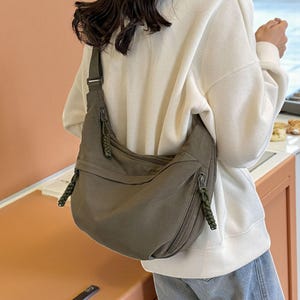 Peut inclure: Sac bandoulière vert olive avec une large bandoulière. Le sac a une forme incurvée avec deux poches zippées à l'avant et des accents tressés décoratifs sur les côtés. Le sac est fait d'un matériau doux et durable.