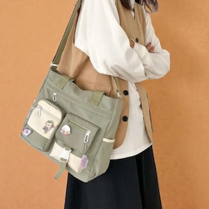 May include: A sage green tote bag with multiple pockets and a shoulder strap. The bag features a small front pocket with a zipper and decorative patches. The bag has a cream-colored accent pocket and a small charm hanging from the zipper.