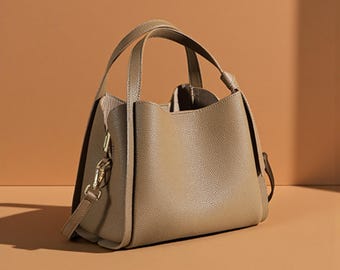 May include: A taupe-colored handbag with a structured design. The bag features two top handles and a detachable shoulder strap. The hardware is gold-toned, and the bag has a pebbled texture. The interior lining is a lighter shade of beige.
