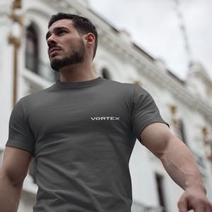 May include: A charcoal gray short-sleeved t-shirt featuring the word VORTEX printed in white capital letters on the left side of the chest. The garment has a classic crew neckline and a plain, solid-colored fabric construction throughout.