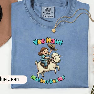 May include: A light blue t-shirt featuring the text "Yee Hawt Mutton Buster" and a cartoon illustration of a girl riding a sheep. A gold necklace with a star pendant is draped across the shirt.