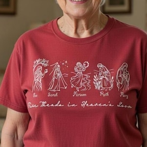 Può includere: T-shirt rossa con grafica bianca raffigurante figure stilizzate e i nomi Eva, Sara, Miriam, Rut e Maria. Il testo "Five Threads in Heaven's Loom" è sotto le figure.