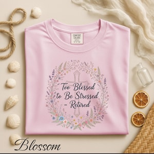 May include: A pale pink t-shirt featuring the text "Too Blessed to Be Stressed - Retired" within a floral wreath and cross design. The word "Blossom" is at the bottom of the shirt.