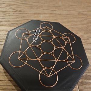 Puede incluir: Un posavasos hexagonal negro con un dise&ntilde;o de Cubo de Metatr&oacute;n de alambre de cobre. Un peque&ntilde;o objeto de cuentas en blanco y negro est&aacute; suspendido sobre el posavasos. El posavasos est&aacute; sobre una superficie de madera.
