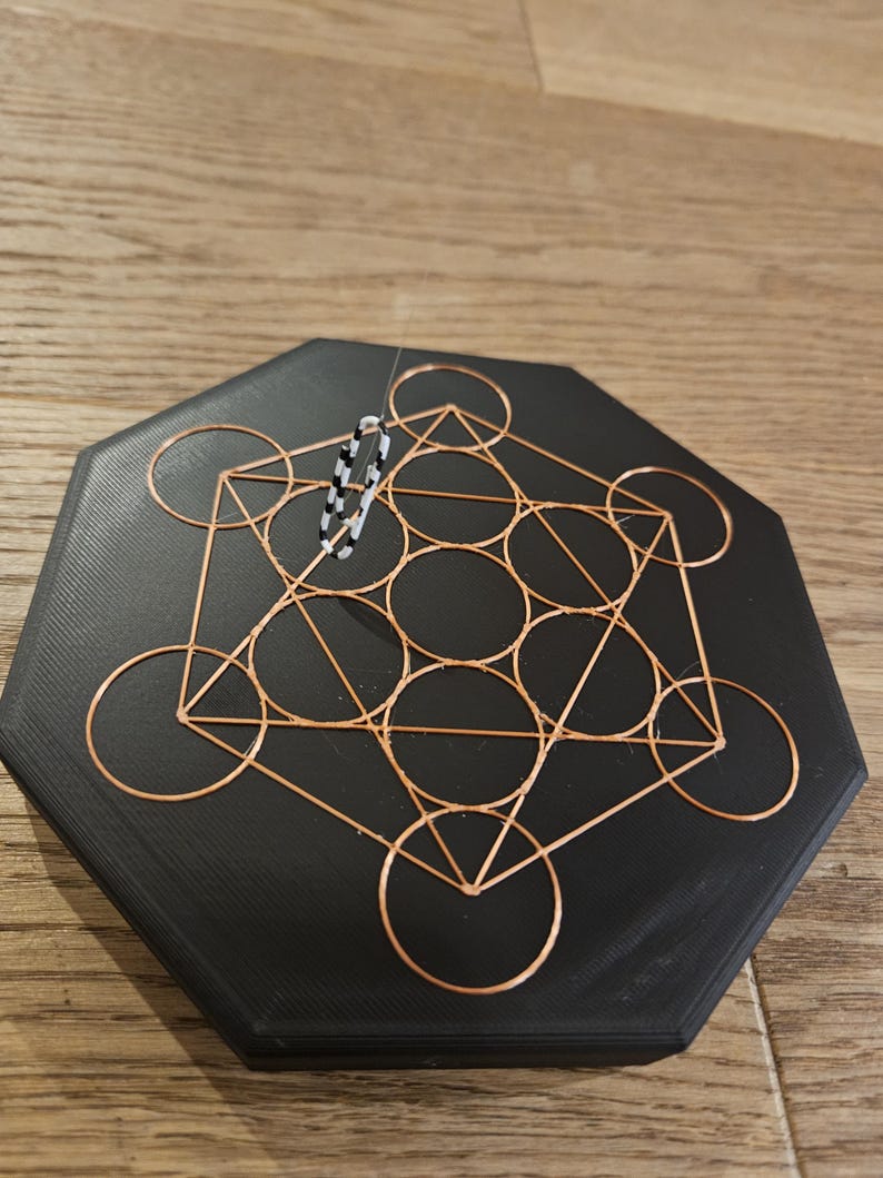 Puede incluir: Un posavasos hexagonal negro con un dise&ntilde;o de Cubo de Metatr&oacute;n de alambre de cobre. Un peque&ntilde;o objeto de cuentas en blanco y negro est&aacute; suspendido sobre el posavasos. El posavasos est&aacute; sobre una superficie de madera.