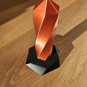 K&ouml;nnte beinhalten: Eine kupferfarbene, geometrische Skulptur mit einem gedrehten Design, die auf einem schwarzen, eckigen Sockel steht. Die Oberfl&auml;che der Skulptur reflektiert das Licht und erzeugt einen Farbverlauf aus Orange- und Braunt&ouml;nen. Der Sockel ist mattschwarz.