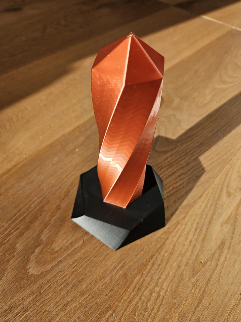 K&ouml;nnte beinhalten: Eine kupferfarbene, geometrische Skulptur mit einem gedrehten Design, die auf einem schwarzen, eckigen Sockel steht. Die Oberfl&auml;che der Skulptur reflektiert das Licht und erzeugt einen Farbverlauf aus Orange- und Braunt&ouml;nen. Der Sockel ist mattschwarz.