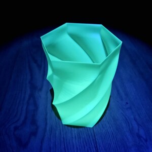 Op de afbeelding: Een lichtgroene, spiraalvormige vaas met een geometrisch ontwerp. De vaas is gemaakt van een glad materiaal en staat op een donkerblauwe houten ondergrond. De vaas heeft een unieke, moderne uitstraling.