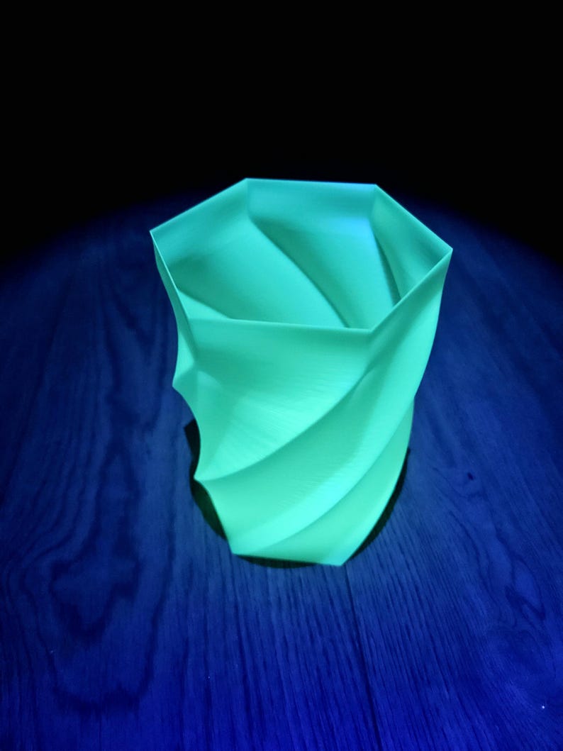 Op de afbeelding: Een lichtgroene, spiraalvormige vaas met een geometrisch ontwerp. De vaas is gemaakt van een glad materiaal en staat op een donkerblauwe houten ondergrond. De vaas heeft een unieke, moderne uitstraling.