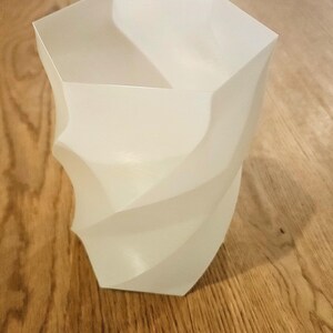 Op de afbeelding: Een witte, geometrische vaas met een gedraaid ontwerp. De vaas heeft een zeshoekige bovenkant en een spiraalvormig, gebogen lichaam. Het object is gemaakt van een glad, ondoorzichtig materiaal en staat op een houten oppervlak.