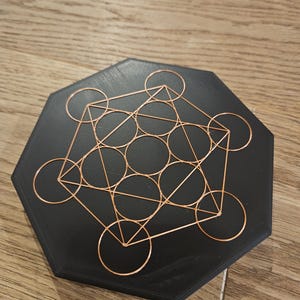 Puede incluir: Un Cubo de Metatr&oacute;n negro hexagonal, un s&iacute;mbolo geom&eacute;trico, con una superposici&oacute;n de alambre de cobre. El alambre forma c&iacute;rculos, tri&aacute;ngulos y l&iacute;neas. El objeto est&aacute; sobre una superficie de madera.