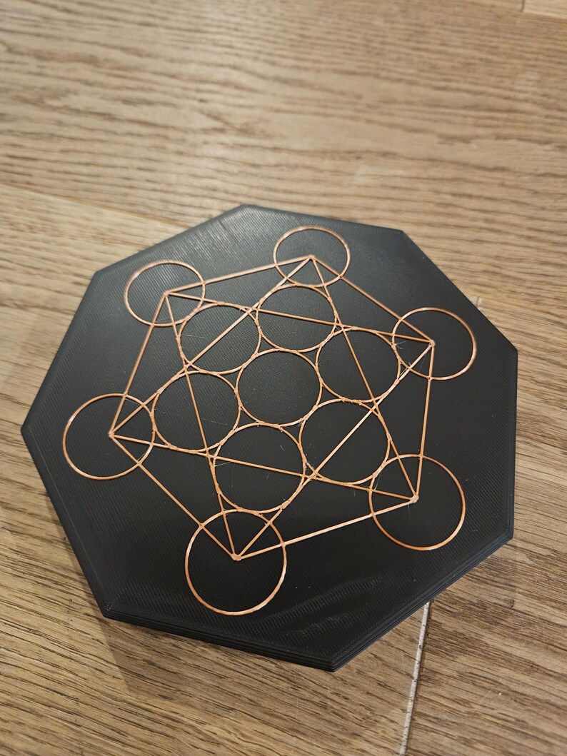 Puede incluir: Un Cubo de Metatr&oacute;n negro hexagonal, un s&iacute;mbolo geom&eacute;trico, con una superposici&oacute;n de alambre de cobre. El alambre forma c&iacute;rculos, tri&aacute;ngulos y l&iacute;neas. El objeto est&aacute; sobre una superficie de madera.