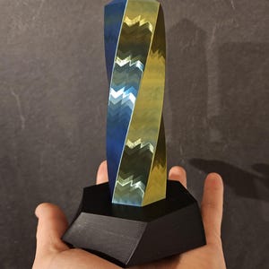 K&ouml;nnte beinhalten: Eine geometrische Skulptur mit verdrehtem Design, mit blauen und goldenen Metalloberfl&auml;chen. Die Skulptur ist auf einem schwarzen, sechseckigen Sockel montiert. Das Kunstwerk wird in einer Hand gehalten, um seine Gr&ouml;&szlig;e und Form zu zeigen.