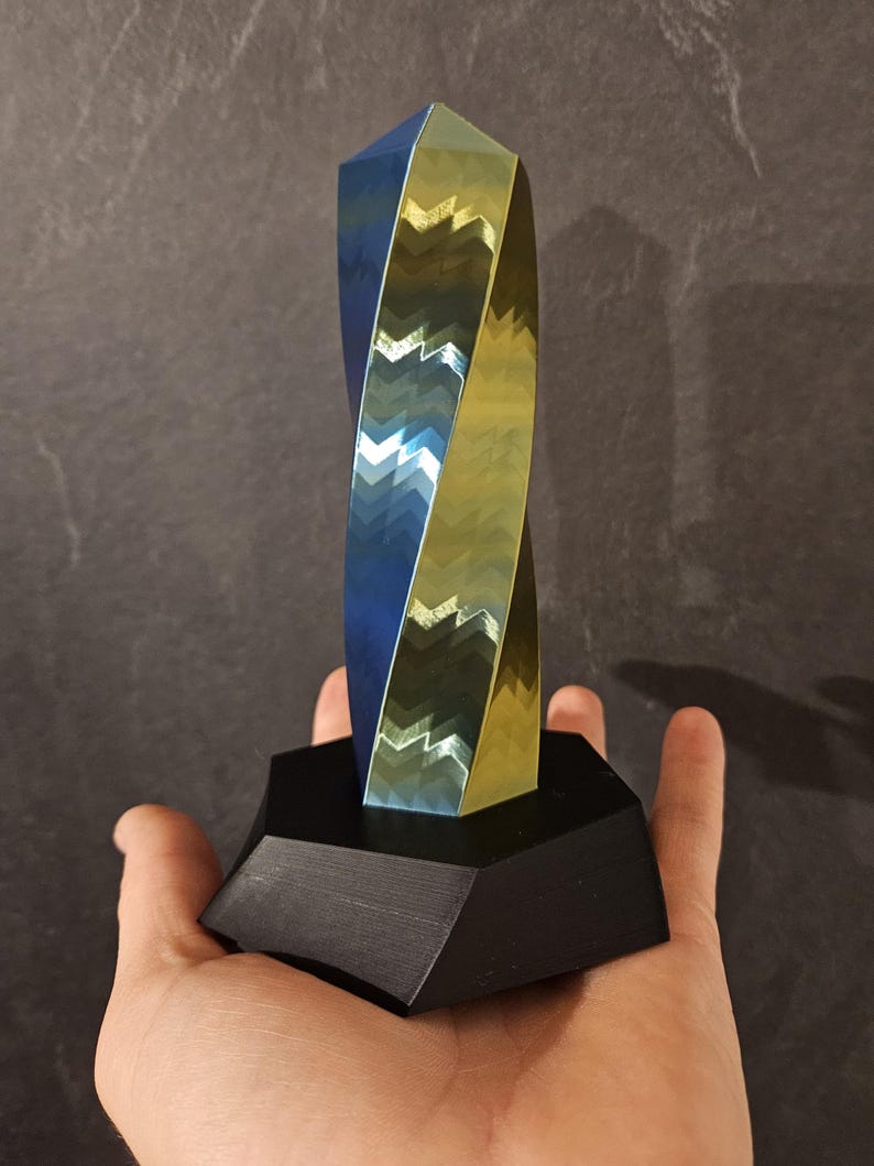 K&ouml;nnte beinhalten: Eine geometrische Skulptur mit verdrehtem Design, mit blauen und goldenen Metalloberfl&auml;chen. Die Skulptur ist auf einem schwarzen, sechseckigen Sockel montiert. Das Kunstwerk wird in einer Hand gehalten, um seine Gr&ouml;&szlig;e und Form zu zeigen.